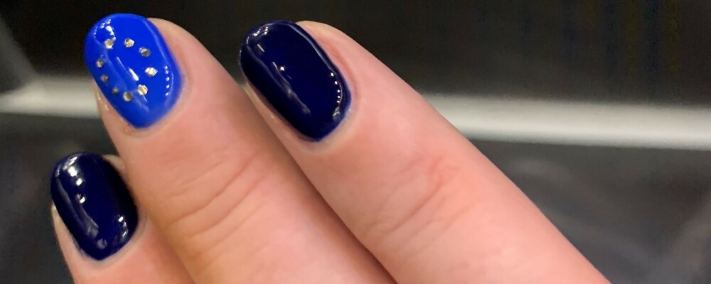 Closeup of some fingers with painted blue fingernails. The middle fingernail has a circle of gold dots painted on it to look like the EU flag.